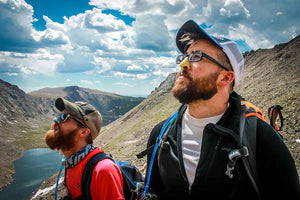 Two male hikers wearing colorful sunscreen gaze into the distance in front of a mountainous backdrop.